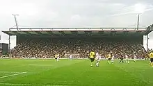 An impressive grandstand behind a goal net, filled with people, viewed from the other end of the stadium.