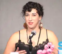 A woman with curly black hair and large disc earrings with scorpions on them speaks into a microphone in front of a gray wall.