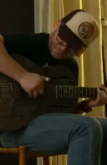 Ross Hammond is playing a resonator guitar. He is seated on a chair, bent over the guitar, wearing a trucker hat and glasses.