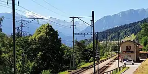 Two-story building with gabled roof situated in mountain valley next to double-track railway line