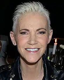 An image of a short-haired woman smiling in to a camera. There is a flame from a candle flickering above her left shoulder.