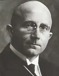 Bald, elderly man with gold-rimmed spectacles, wing collar, and serious expression