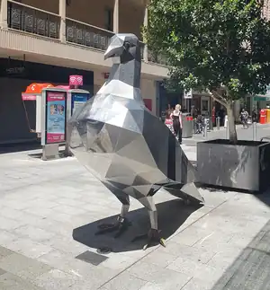 Showing a 2 metre tall silver stainless steel pigeon sculpture, shining in the afternoon Australian sun. Standing upon grey tiles with a tag around its ankle and standing up.