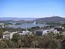 A group of multi-story office buildings. A lake, mountains and a jet of water are visible in the background.