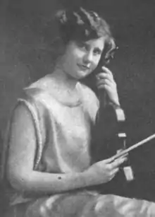 A young white woman, seated, holding a violin upright in her lap, and a bow in her right hand. She has short wavy hair, and is wearing a light-colored satiny dress with no sleeves.