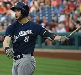 A man in a navy baseball uniform with "Brewers" across the chest, a navy batting helmet, and gray pants holding a baseball bat