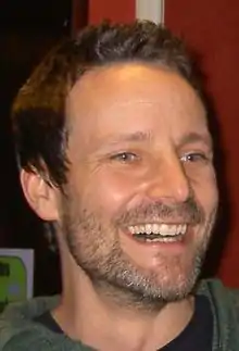 A head shot of a shorthaired man with facial hair, is grinning at an audience.