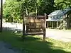 A brown wooden sign with yellow letters reading "Sand Bridge State Park Picnic Area Pennsylvania Department of Conservation and Natural Resources" in front of a highway and pale green two story frame house