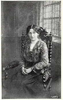 A black and white photograph of a middle-aged woman dressed in formal Edwardian clothing. She is sitting on an old chair in a large room.