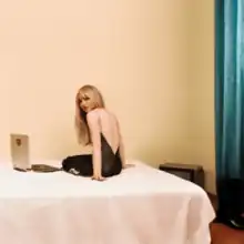 Sabrina Carpenter wearing a bareback black dress and sitting on a hotel bed, with a laptop next to her.