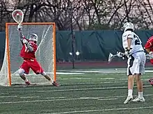 A man wearing a red jersey and shorts with a white helmet and gloves holds a white stick with a large meshed area on one end just above a square of metal rods with netting behind it. On the right another man, wearing white jersey and green trim but otherwise similarly attired to the first, holds a similar stick with a much smaller head at his right.