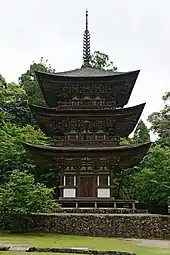 A brown wooden pagoda on a stone base. In the background: trees with green foliage.