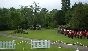 Red coated band members stand in formation on the lawn, playing.