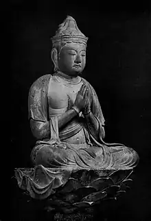 Three-quarter view of a cross-legged seated deity on a pedestal with hands joined as in prayer in front of the body.