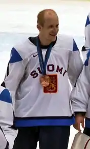 A Caucasian ice hockey player dressed in a blue jersey with green and white trim, as well as a blue helmet. He stands relaxed on the ice, holding his hockey stick in one hand.