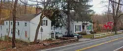 Three houses seen from an elevated position across a road. The two on the left are white, with the leftmost facing the road side on. Between it and the house next to it is a driveway with two cars and a tall evergreen tree. Further down the road, at the right, the house is red.