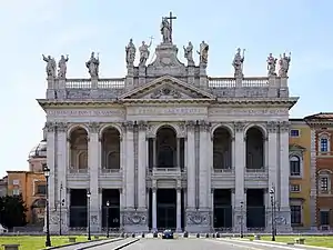 Ornate facade of the basilica at night with columns, main door, and statues of the twelve Apostles on the roofline, with a Latin inscription below them