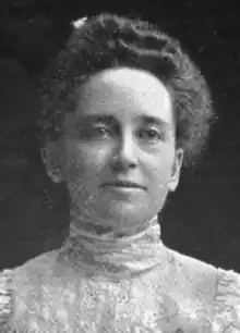 A white woman with hair in a bouffant updo, wearing a high-collared white lacy blouse or dress