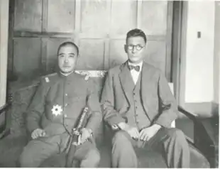 A black and white photograph of two seated men: one wearing a Japanese officer's uniform, the other a formal suit including a bow tie