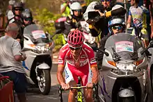 A road racing cyclist in his early thirties, wearing a predominantly red jersey with white trim and a red helmet. A motorcycle bearing a cameraman follows behind him, with a further caravan and other spectators in the background.