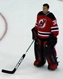 An ice hockey player standing, looking to the left of the camera. He is wearing a red and black uniform and is holding his hockey stick out in front.