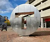 A large metal disk with "T" cut out from the middle, in front of a parking garage