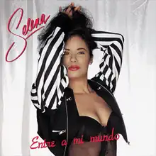 Cover album for Entre a Mi Mundo. Selena is featured in a close-up portrait, posed with her hands entangled in her hair. She wears an unzipped short black jacket with black-and-white patterns adorned on its sleeve, exposing her midriff in front of a clear background. The album title, Entre a Mi Mundo, is written in red cursive at the bottom of the artwork, while Selena's name is presented in red cursive letters in the top left corner.