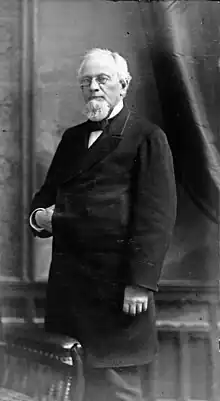 A black and white photograph of a standing elderly bespectacled man wearing a bow tie and dark frock coat with his right hand in between the seam. His body is angled to the right while he looks straight at the camera.