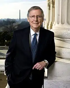 A man in his seventies with graying hair and glasses leaning against a column and wearing a black suit, white shirt, and red tie