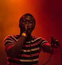 The image is cropped to show the upper half of Mubarik Adams on a dark orange background. Mubarik has short hair and is wearing a horizontally striped T-shirt and a yellow wrist band. He is holding a mic to his mouth with his right hand while holding the mic's cord in his outstretched left hand. His eyes are closed and his left hand is pointing upward.