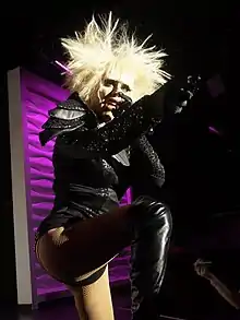 Female-presenting individual on a stage, with blonde hair and wearing a black outfit.