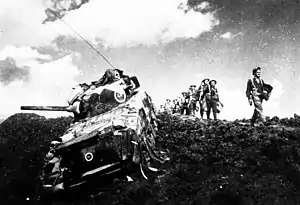 A tank of World War II vintage, with some foot soldiers marching alongside