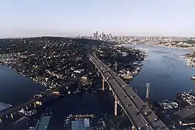  A freeway with a smaller bridge next to it spanning a waterway to a wooded but still urbanized hillside with a city's skyline in the distance