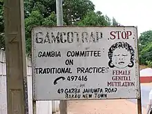 A fading sign that says: GAMCOTRAP - Gambia Committee on Traditional Pratices. 49 Garba Jahumpa Road, Bakau New Town. There is a phone next to the numbers 497416. On the ride side of the sign, there is a drawing of a crying woman. Above her, it says STOP; below, it says FEMALE GENITAL MUTILATION. This text is in red.