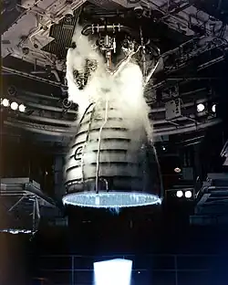 A rocket engine firing. A blue flame is projecting from a bell-shaped nozzle with several pipes wrapped around it. The top of the nozzle is attached to a complex collection of plumbing, with the whole assembly covered in steam and hanging from a ceiling-mounted attachment point. Various pieces of transient hardware are visible in the background.