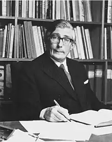 Photograph of a man in a formal suit, writing at a desk.