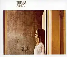 A woman with dark blonde hair, facing left, stands against a white door behind her. Her head is turned slightly toward the viewer. The background is a wooden wall. Surrounding this picture is a white border with a thicker top line. On this line, halfway between the left edge and center, is written "TRAVIS", and below it, "SING".