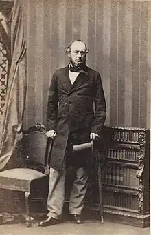 Sir John Duckworth standing between a chair to the left and low bookcase to the right, holding a top hat in his left hand