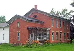 A brick house with pointed roof and chimney. Some of its windows are missing and a wooden section in front is in poor repair. A white wooden wing projects from the rear at the left of the image.