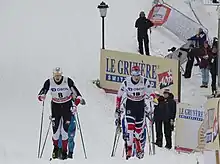 Two lines of cross country skiers are seen from a long ways away, but zoomed in. The lines are skiing directly towards the camera, led by two men from the same team. A few spectators can be seen off to the side. The blank snow carpets everything