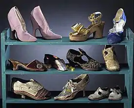 Three blue shelves on a black background. The top shelf displays a pair of pink high heels, a wedge sandal and a dance heel shoe. The middle shelf displays a variety of low-heeled, slipper-type shoes. The bottom shelf displays two antique heeled shoes and a pair of kid's shoes.