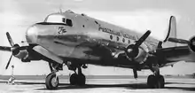 Close-up, in black-and-white, of a silver-bodied four-engined propeller aircraft standing on an apron at an airport