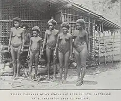 Black-and-white photograph of five nearly naked young women and girls standing in front of a hut; a sixth is visible in the background