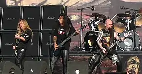 Three men on a stage, all holding guitars. All three are wearing black clothing, and audio equipment can be seen both in front of and behind them.