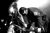 Black and white image of three men wearing jackets and masks over their faces. The one in the forefront is bent over, holding a guitar.