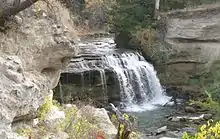 Small river going off rocky ledge over overhang, low cliffs alongside