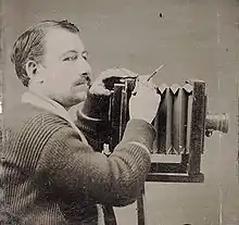 Dark-haired man with small moustache; operating large bellows camera