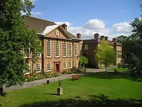  A large brick building is shown, left, with long upper windows above which is classical pediment which includes a round window. In front of the building is an area of grass and trees.