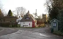 Village centre with two thatched single story cottages with white walls, a red phone box, a red post box and a greenish wooden bus stop shelter with a pitched roof