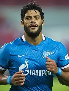 Man with dark skin and short black hair, wearing a football kit composed of a yellow shirt with green collar and blue shorts, running in front a filled stadium stand.